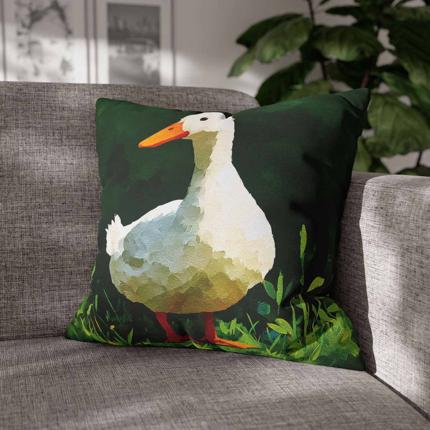 A square throw pillow featuring a stylized low-poly illustration of a white duck with an orange beak and red legs on a dark green background, resting on a gray fabric sofa with a houseplant in the background.