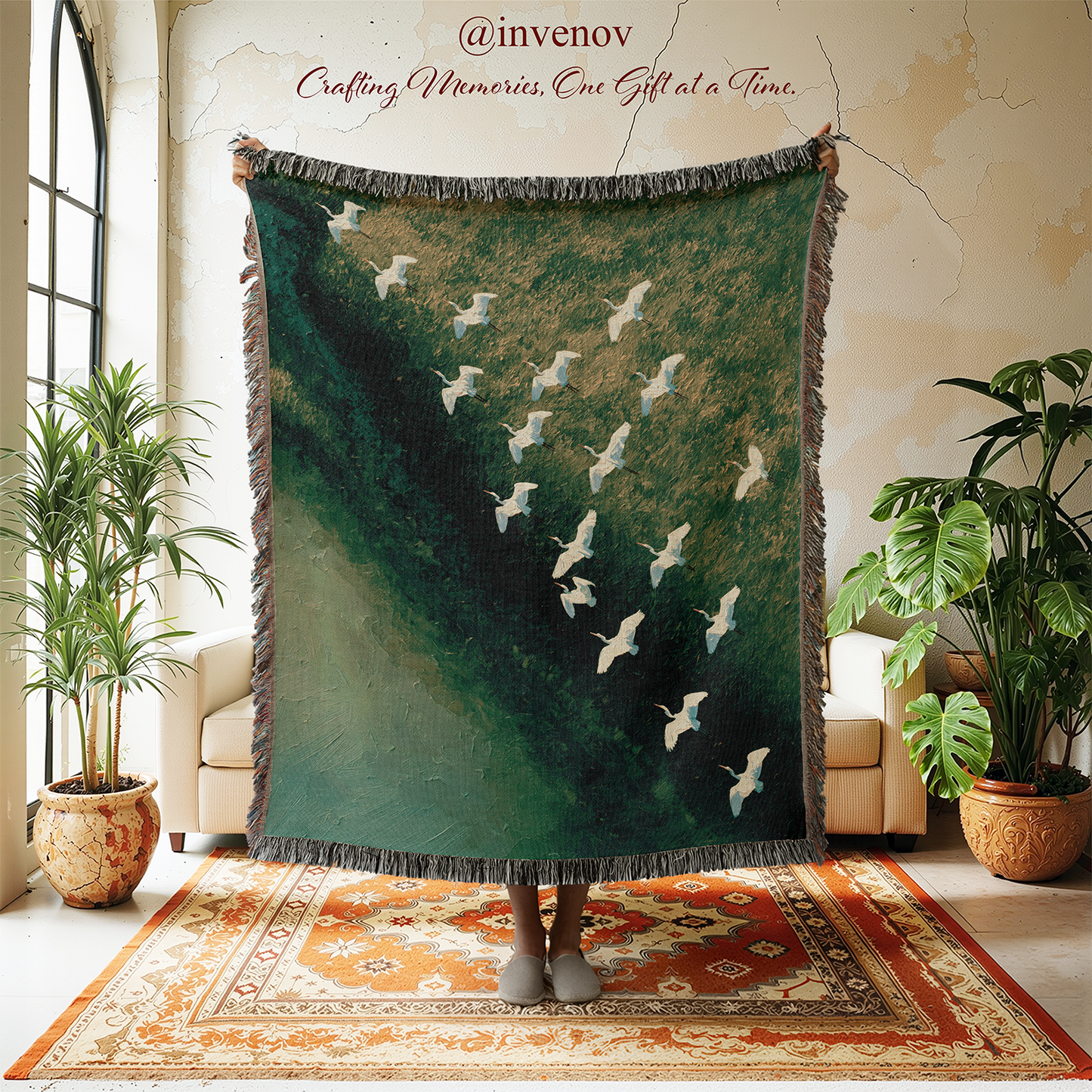 A large woven throw blanket with fringe is held up in a styled room. The blanket features a dark green and teal landscape design with white cranes flying diagonally across it.