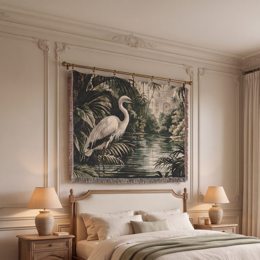A large woven tapestry featuring a white heron in a tropical jungle by a lake hangs on a bedroom wall above a bed with two lit bedside lamps.
