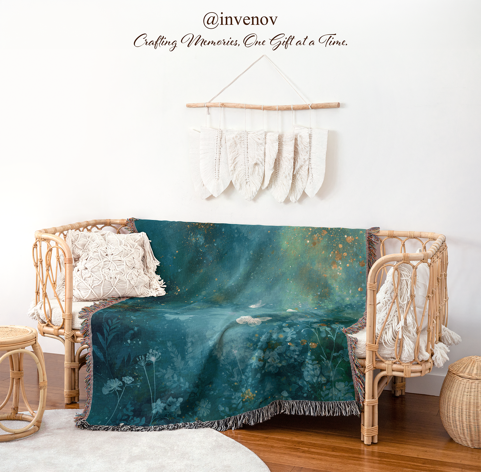 A teal and blue botanical throw blanket with white floral details and a fringed edge is draped over a rattan daybed in a styled room. The scene includes a white wall, macrame wall hanging, side table, rug, and woven stool