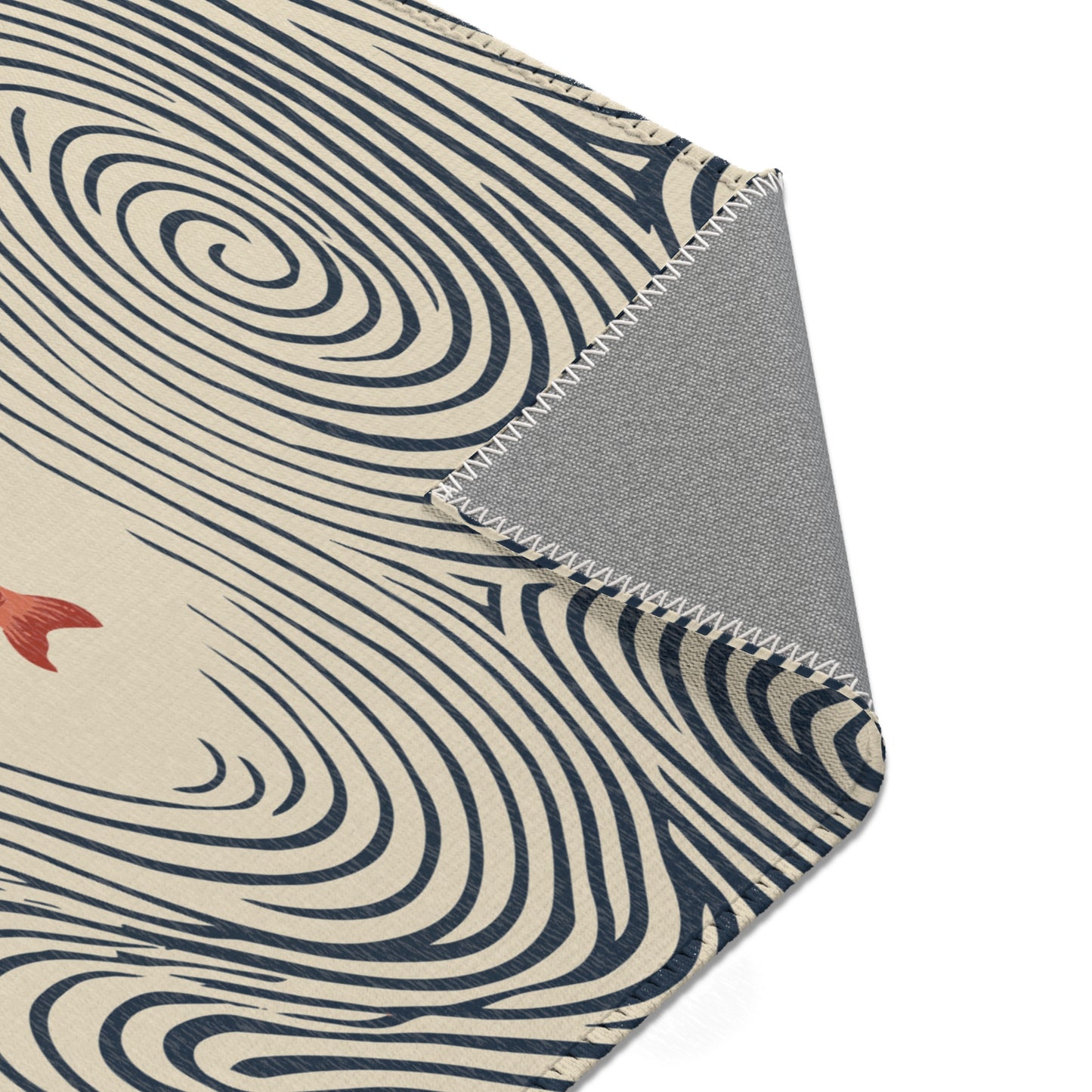 A close-up of an area rug corner showing a cream base with dark navy swirling wave-like lines and a small red fish motif, with a folded gray underside visible along a zigzag stitched edge on a white background.
