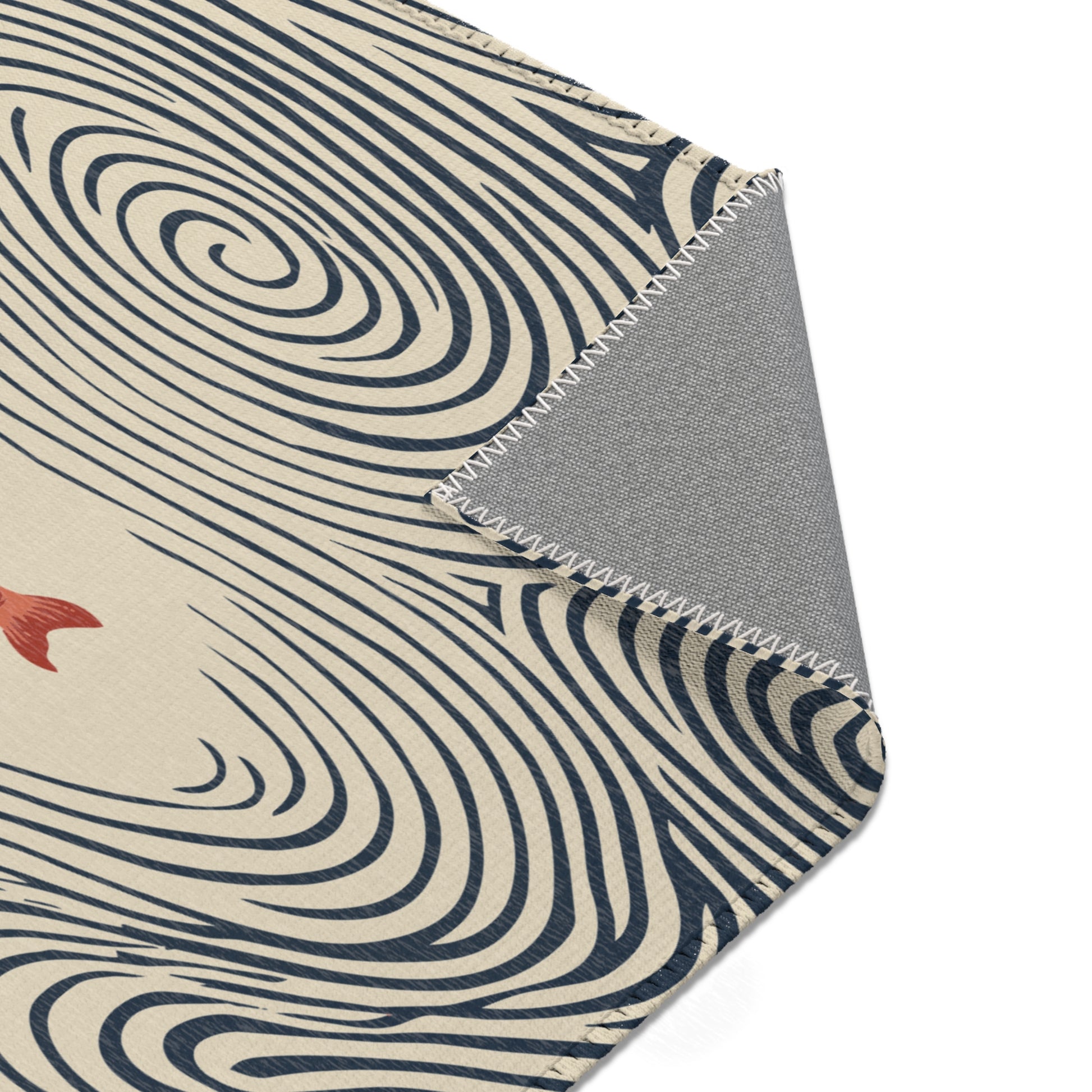 A close-up of an area rug corner showing a cream base with dark navy swirling wave-like lines and a small red fish motif, with a folded gray underside visible along a zigzag stitched edge on a white background.