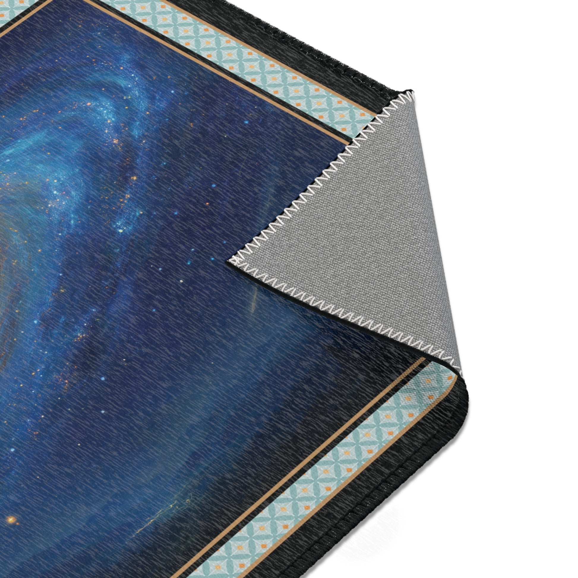 A close-up of a rectangular mat or rug with a dark blue galaxy print, a patterned border in light blue and beige, and a folded corner showing a gray textured underside, set against a plain white background.
