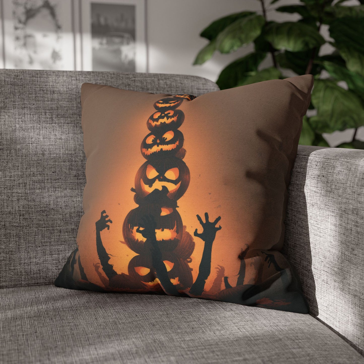 A square throw pillow with a Halloween design featuring a stacked stack of jack-o'-lanterns and zombie hands, resting on a gray textured sofa in a living room setting.
