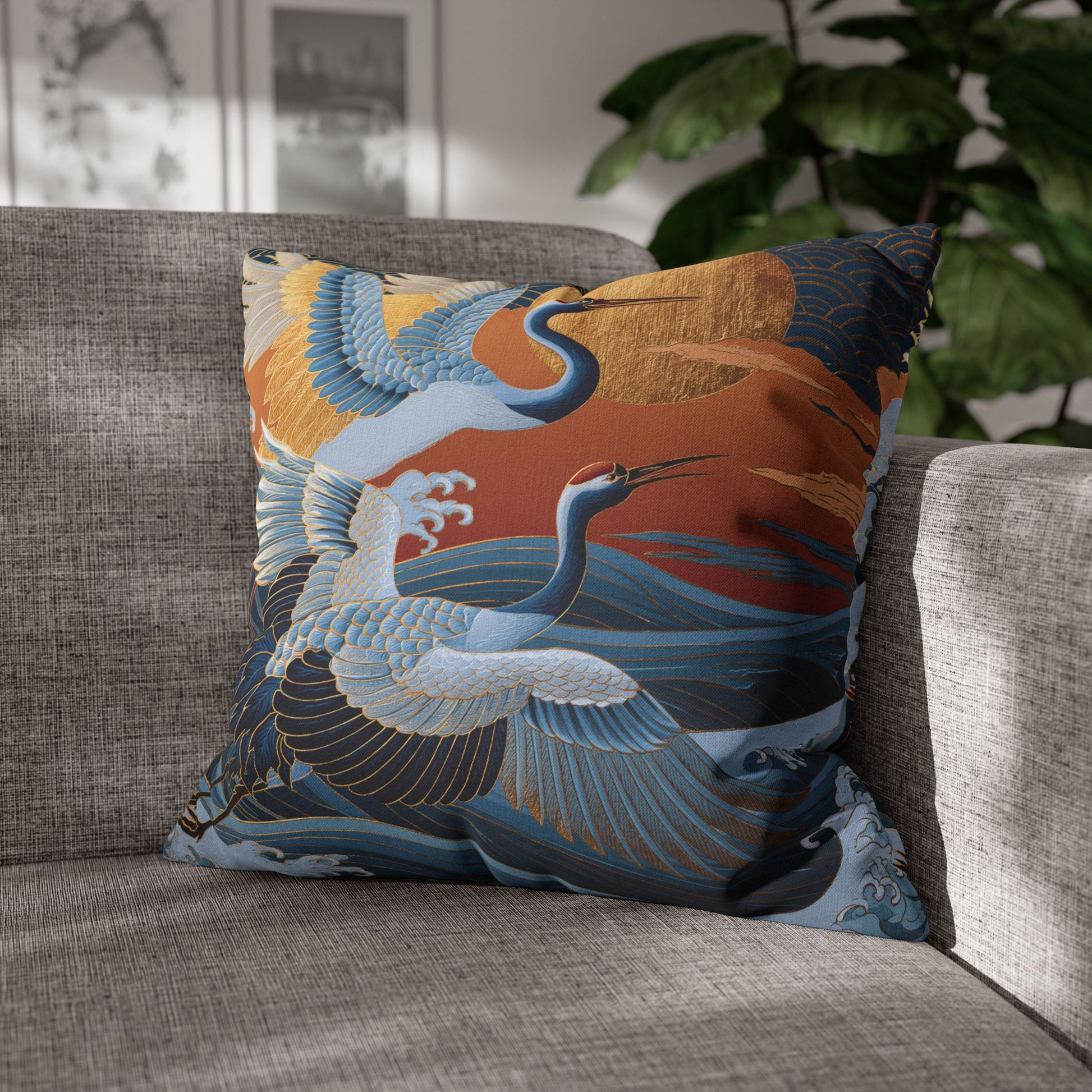 A square decorative throw pillow featuring a Japanese-style crane print in blue, white, orange, and gold, resting on a gray textured sofa with a blurred indoor background.