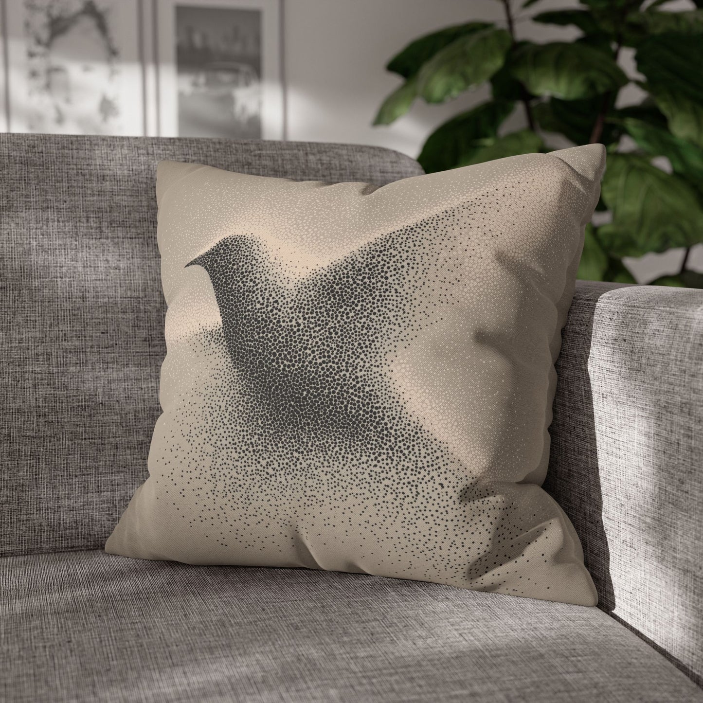 A square throw pillow with a black bird silhouette made of tiny dots on a beige background, resting on a gray textured sofa in a sunlit room with a green plant in the background.