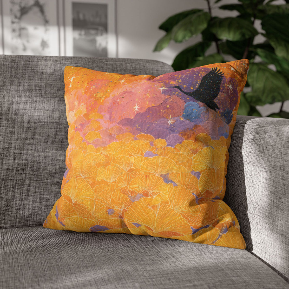 A square decorative throw pillow with a vibrant orange, yellow, and purple ginkgo leaf pattern and a black bird silhouette, resting on a gray fabric sofa in a living room setting.