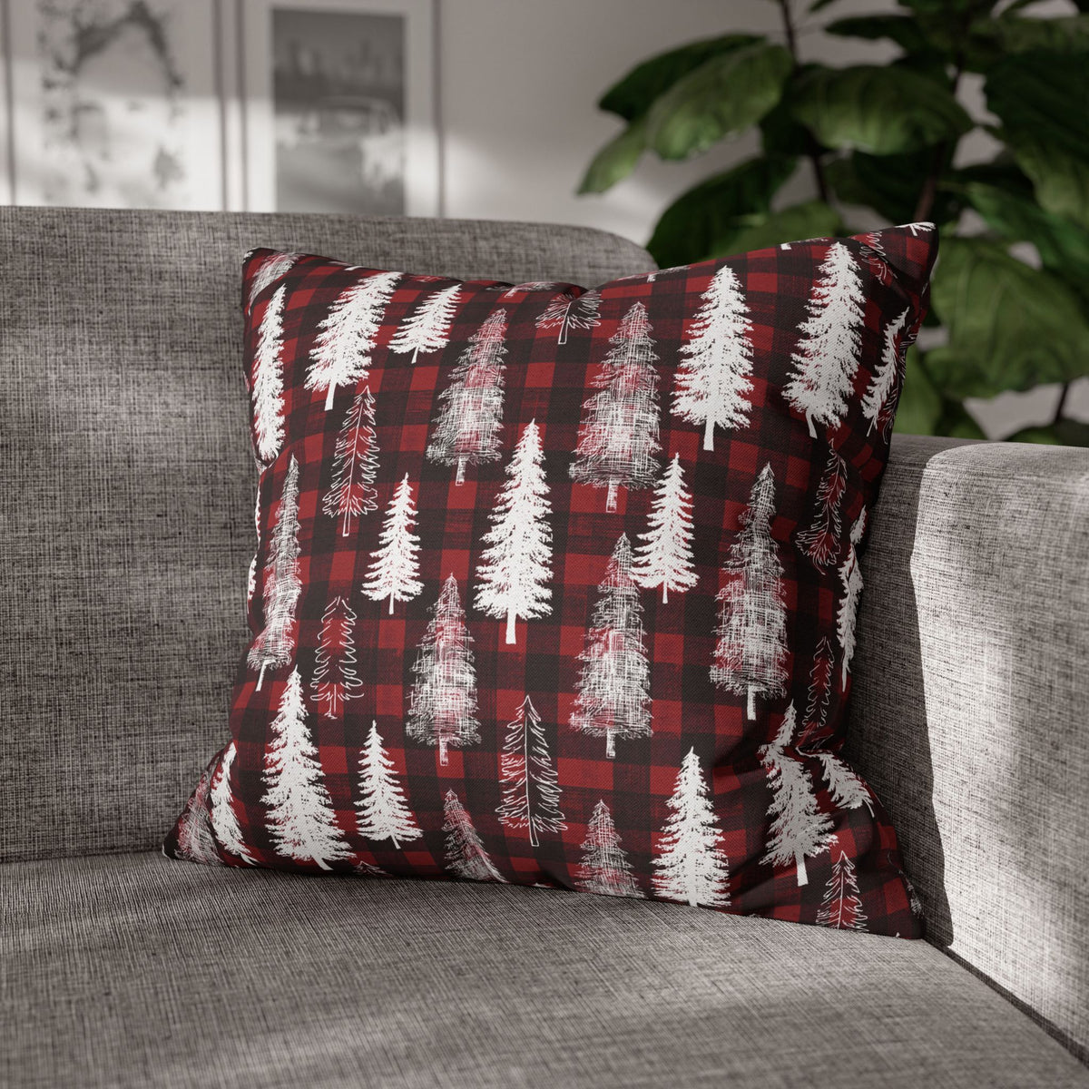 A square throw pillow with a red and black buffalo plaid pattern overlaid with white pine tree silhouettes, resting on a gray fabric sofa with a blurred indoor background.