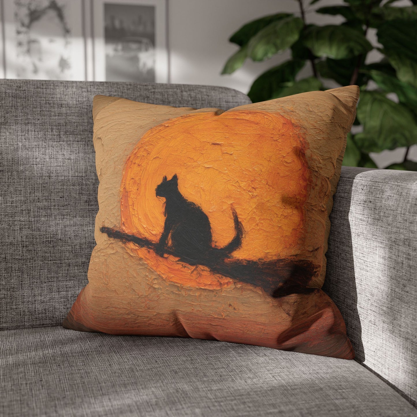 A square throw pillow with a textured painting of a black cat silhouette against a large orange sun, resting on a gray fabric sofa with a green plant in the background.