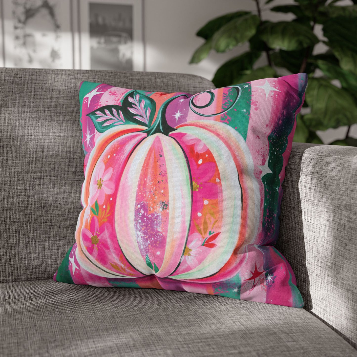 A square throw pillow with a colorful, artistic pumpkin design resting on a gray fabric sofa in a living room setting with a plant in the background.