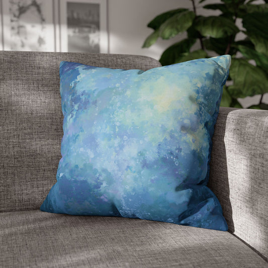 A square throw pillow with an abstract watercolor-like pattern in shades of blue, teal, and light yellow, resting on a gray textured sofa with a green plant in the background.
