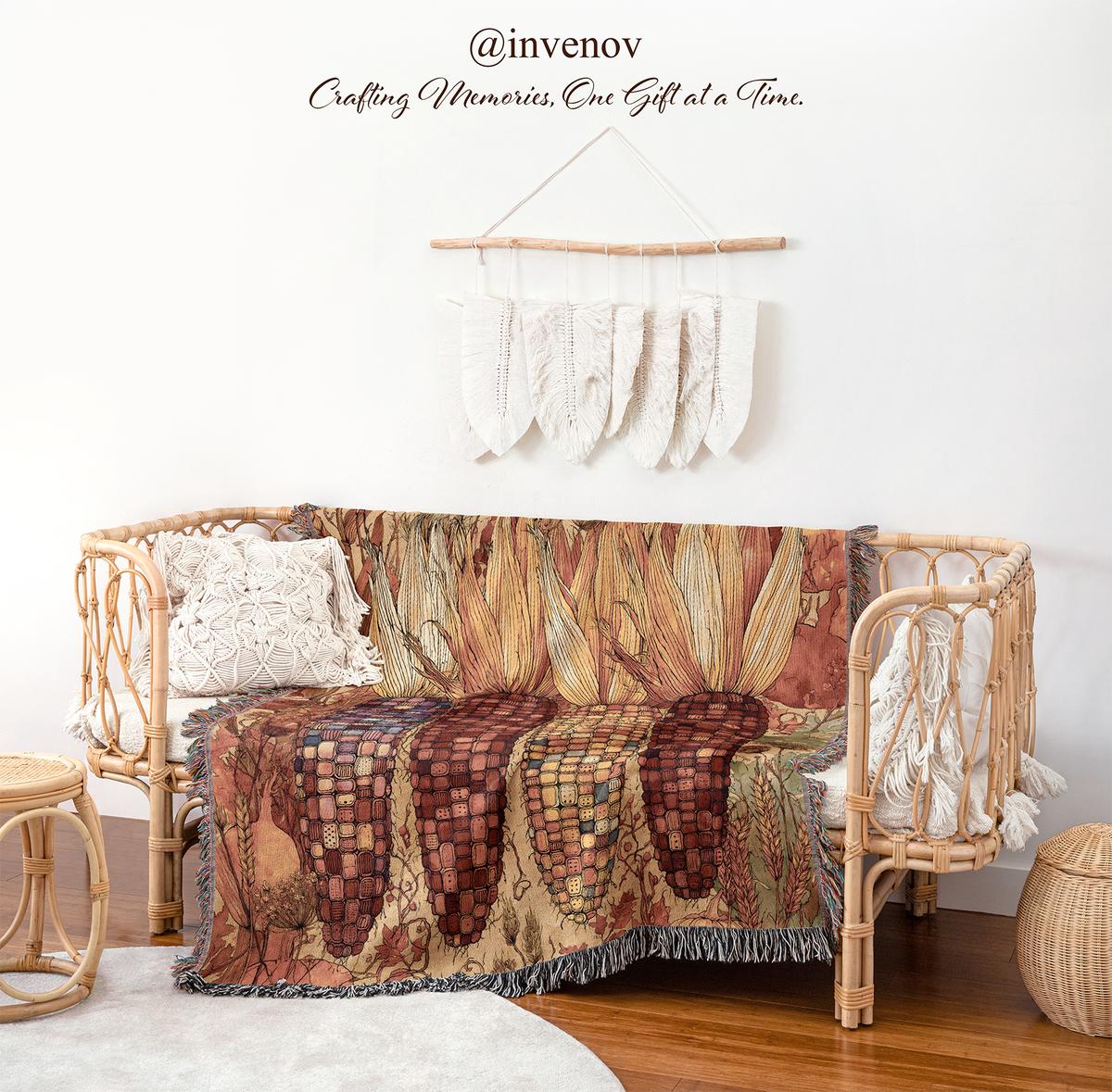 A woven rattan daybed styled in a bohemian room setting, draped with a detailed botanical patterned throw blanket. The scene includes a white wall with macrame wall hanging, wooden floor, a small woven stool, and a round wicker basket.