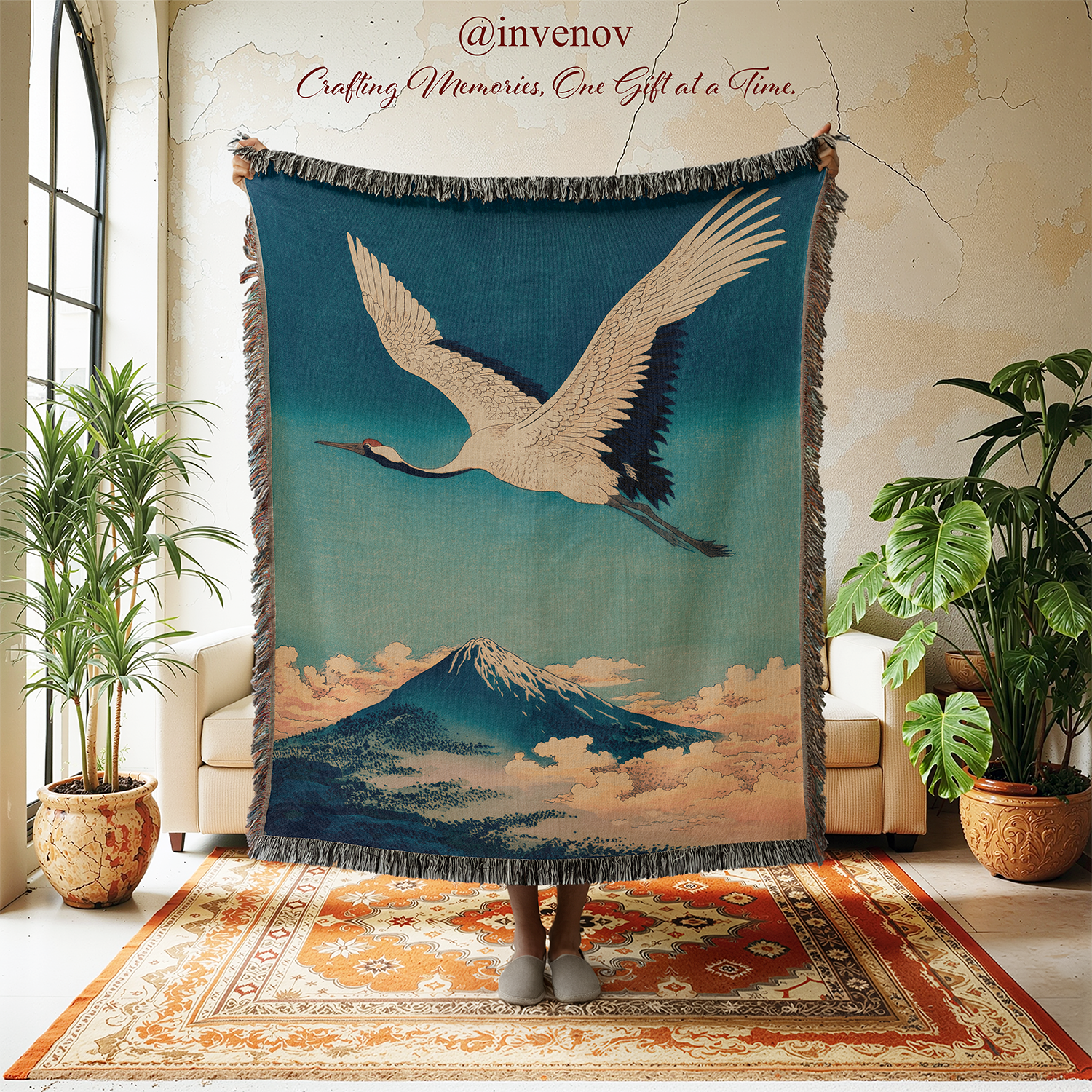 Decorative blanket with crane and mountain design held up in a room with plants and furniture.