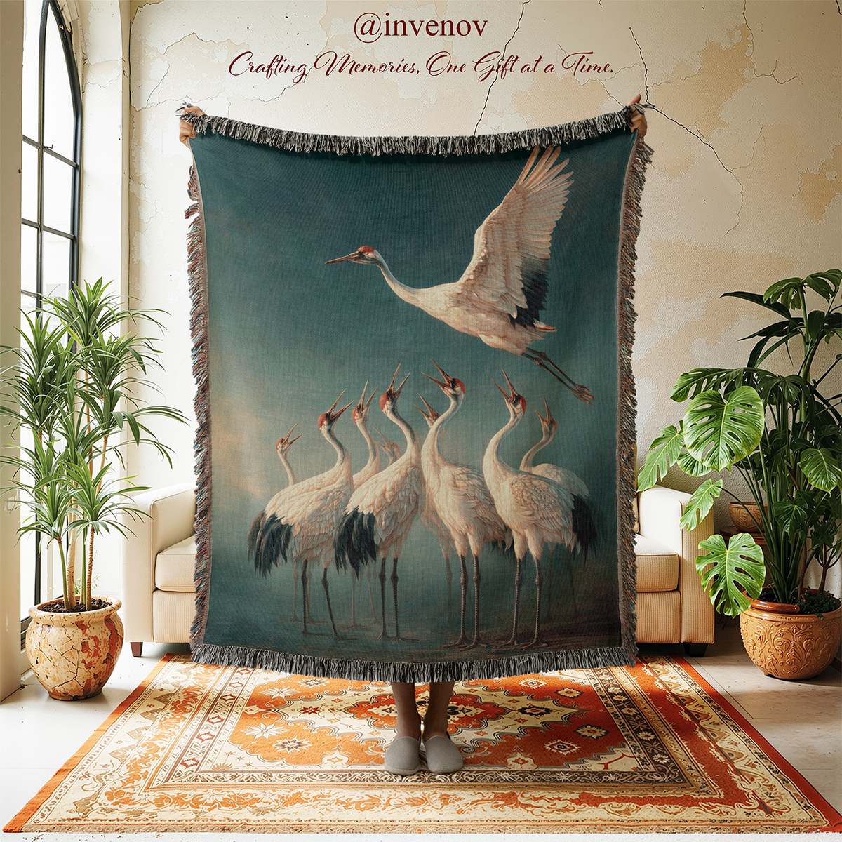 A large woven throw blanket with fringe is held up in a styled room. The blanket features a detailed print of several cranes on a muted teal-blue background, with one crane flying above a group.