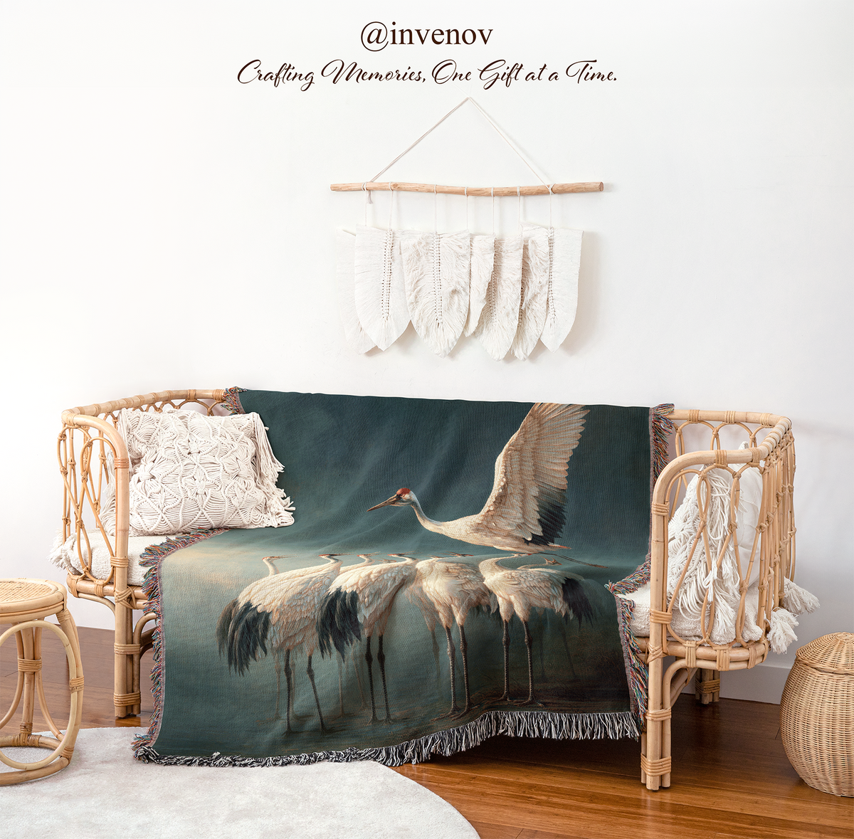 A large woven throw blanket featuring a painting of white cranes on a dark teal background, draped over a rattan daybed in a styled room setting with a macrame wall hanging and wooden floor.