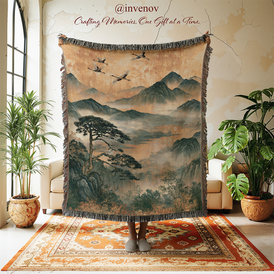 A large fringed tapestry featuring an East Asian style landscape painting with mountains, trees, and birds, held up by a person standing on a patterned rug in a styled room with potted plants.