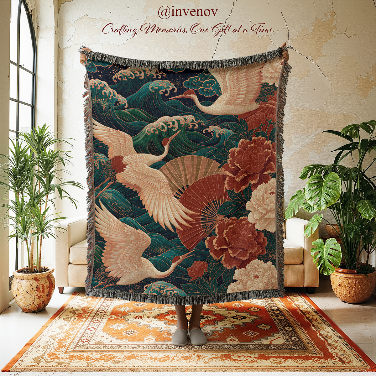 A large woven tapestry featuring cranes, waves, and flowers is held up in a styled room with potted plants, a rug, and cream-colored chairs. The tapestry has a dark blue background with beige cranes, red and pink floral motifs, and green waves, and is shown with a person's feet visible at the bottom.