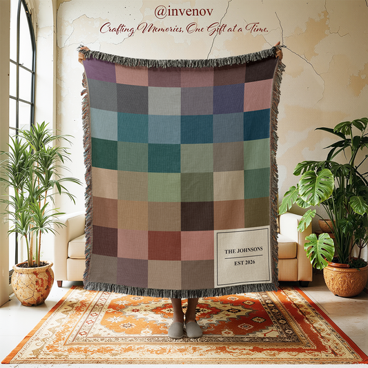 A person holds up a large woven throw blanket with a patchwork design in muted earthy tones, standing on a patterned rug in a styled room with plants and cream chairs. The blanket has fringe around the edges
