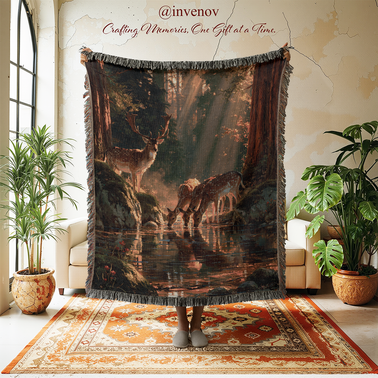 A large fringed throw blanket featuring a forest scene with deer and a reflective waterway, held up in a styled room with plants, cream chairs, and a patterned rug. The blanket has dark gray fringe and a dark, moody print with warm brown and orange tones.