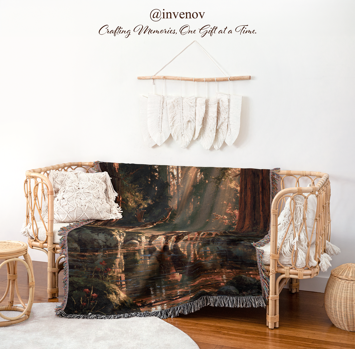 A woven rattan daybed styled in a boho room setting, draped with a dark forest landscape throw blanket featuring a lake and trees. The scene includes white textured pillows, a small woven stool, a round basket, and a macrame wall hanging on a white wall with the brand name @invenov above.