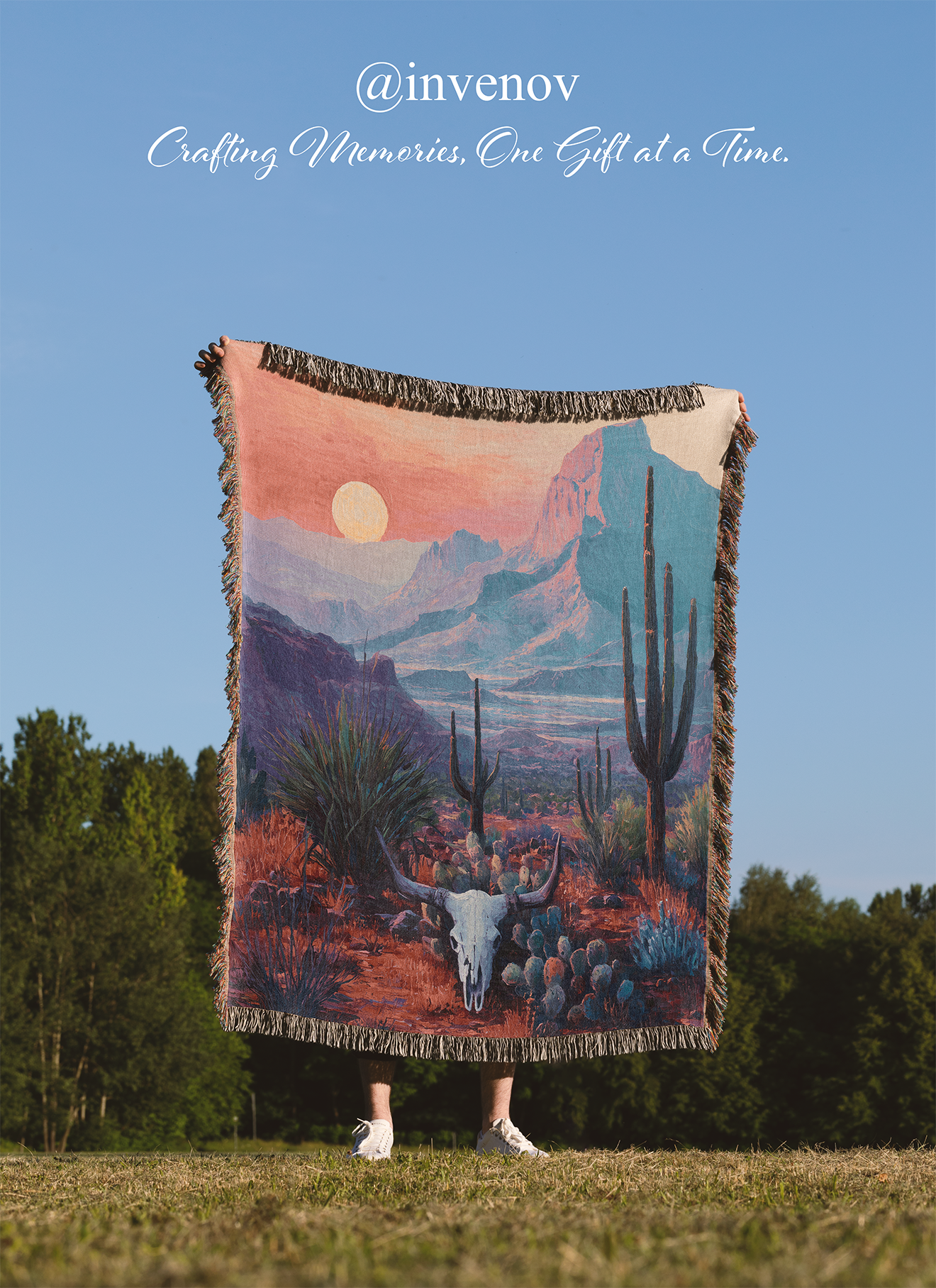 Person holding a desert landscape blanket with mountains and cacti against a blue sky.