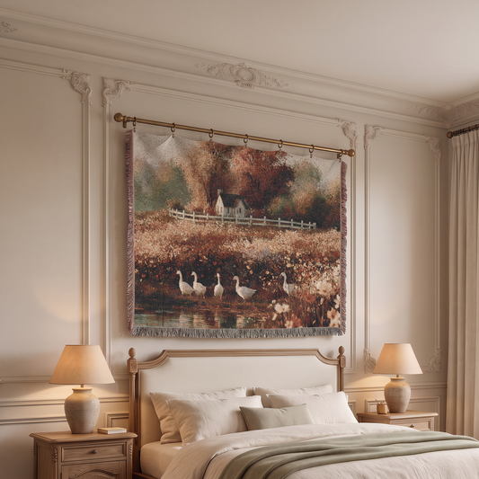 A large woven wall tapestry featuring a rustic autumn landscape with geese by a pond, hung above a bed in a styled bedroom with cream walls, lamps, and wooden nightstands.