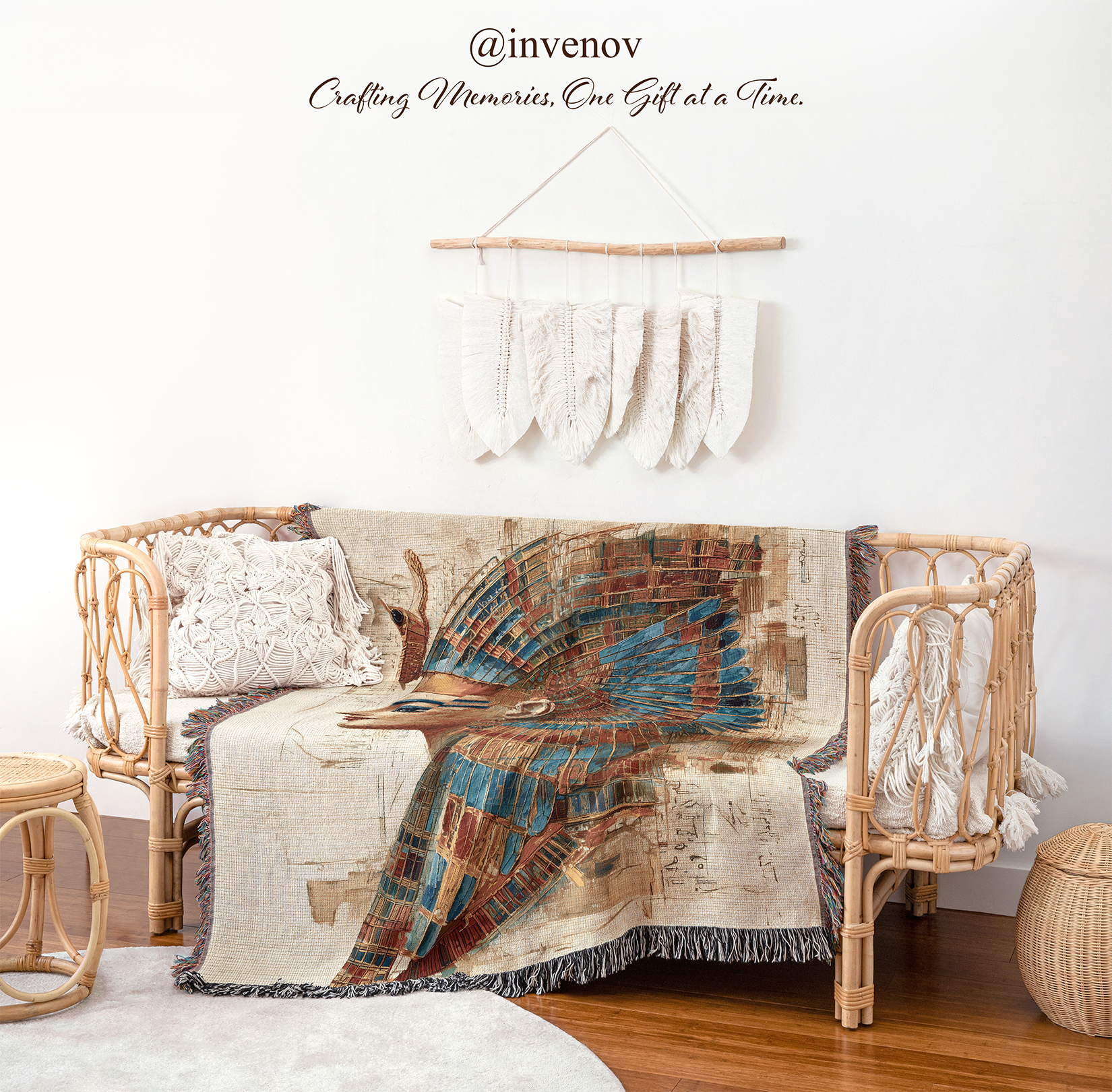 A woven rattan daybed styled in a bohemian room setting, draped with a large abstract peacock-patterned throw blanket. The scene includes a matching woven pillow, a small woven side table, a round floor cushion, and a white wall hanging with tassels.