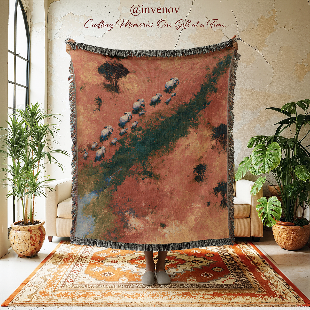 A person holds up a large woven throw blanket with fringe in a styled room. The blanket features an artistic landscape print with a dusty rose and terracotta background, dark green river-like lines, and elephants walking along a path.