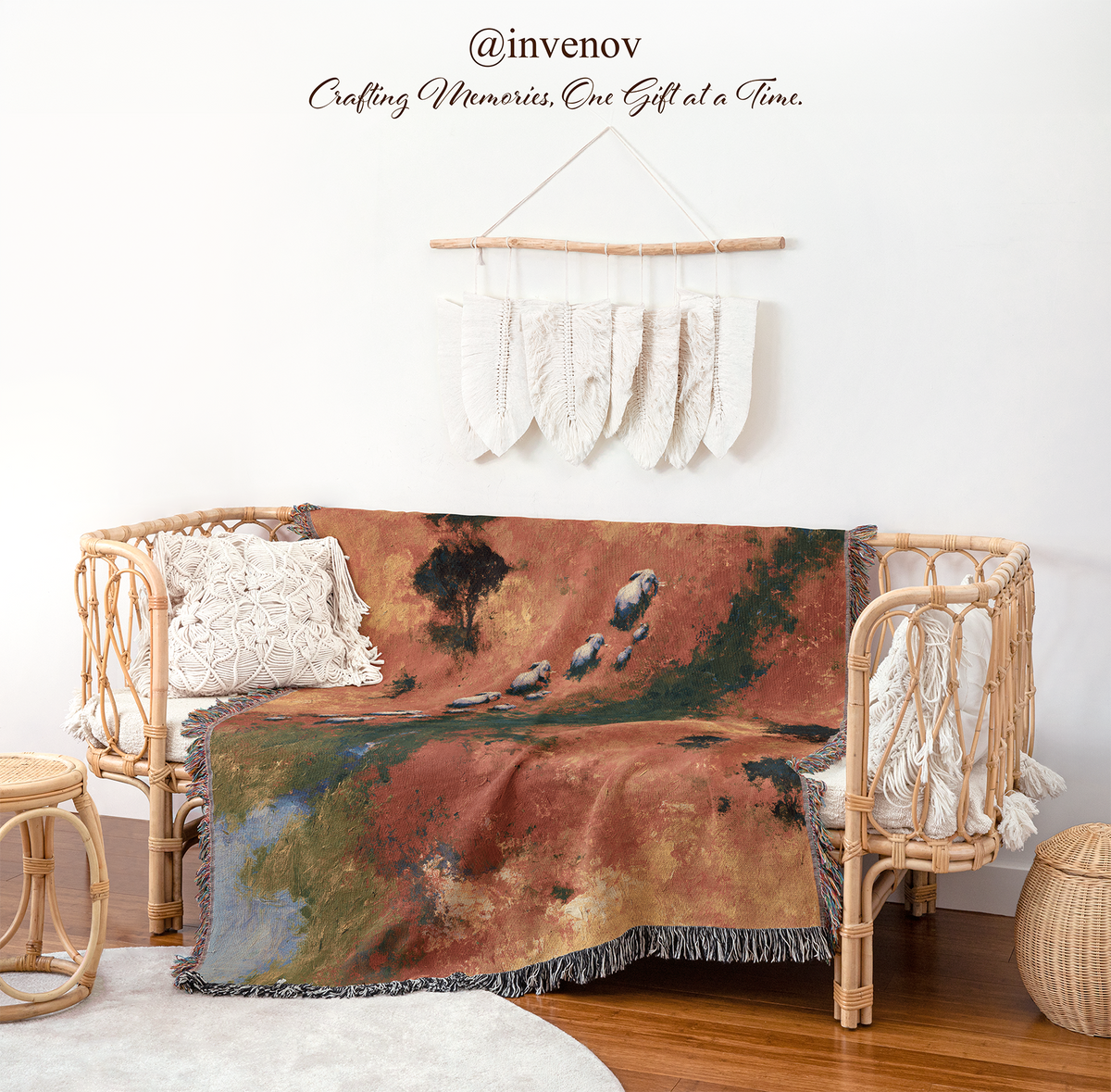 A woven rattan daybed styled in a bohemian room setting, draped with a large abstract landscape throw blanket. The blanket features a warm reddish-brown and orange background with green and blue accents.