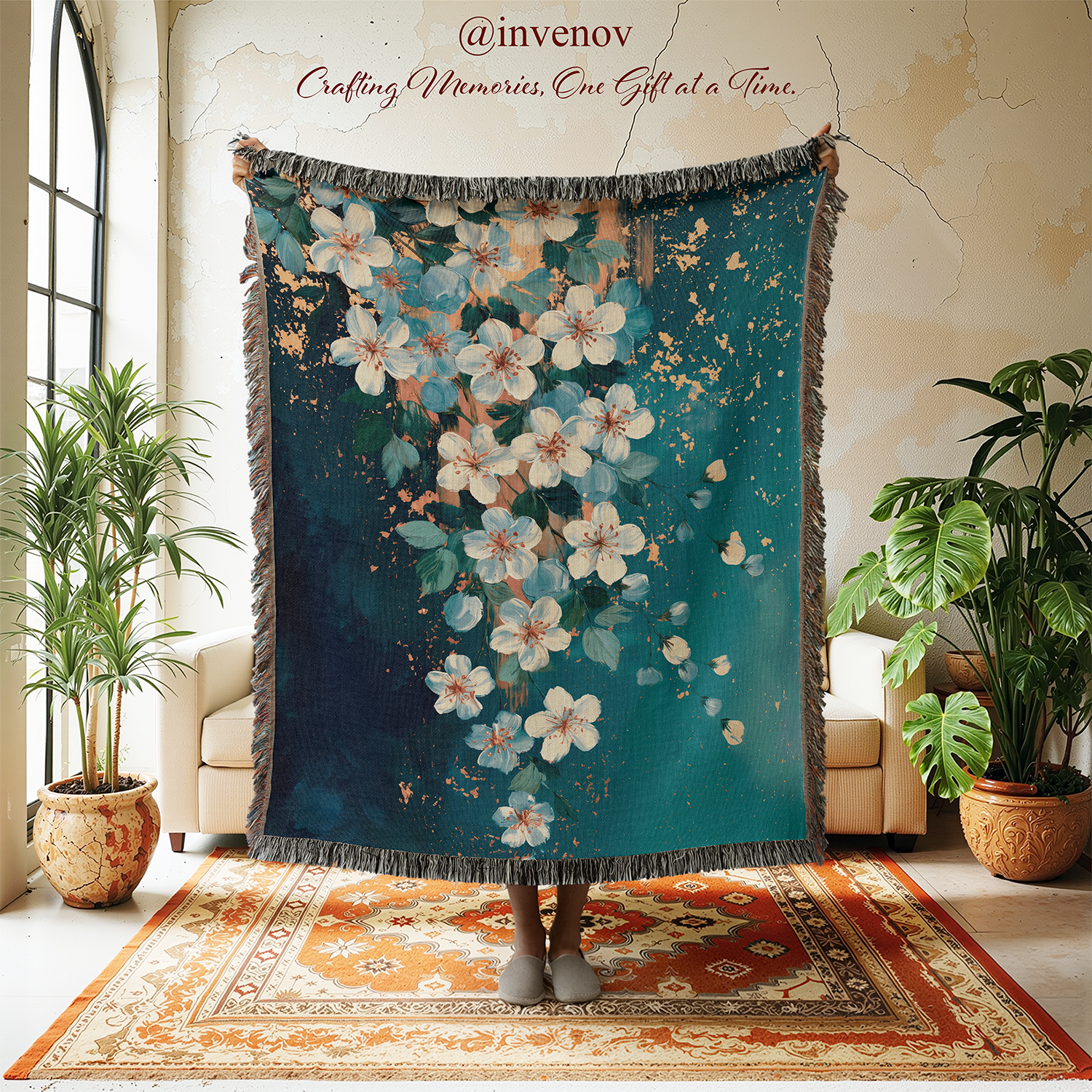 A large woven throw blanket with fringe edges is held up in a room. The blanket has a dark teal-blue background with a floral pattern of white blossoms and small gold accents.