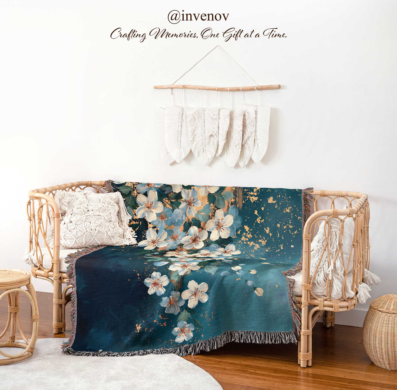 A teal and navy floral throw blanket with white blossoms and gold accents is draped over a rattan daybed in a styled room. The scene includes a white wall, decorative hanging textile, pillows, wood floor, and a small woven stool.