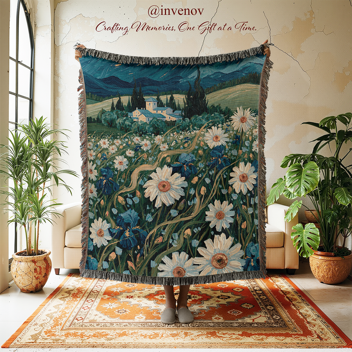 A large woven tapestry featuring a landscape painting of a daisy field and cypress trees, held up in a styled room with plants, chairs, and a rug. The tapestry has gray fringe and the text '@invenov Crafting Memories, One Gift at a Time.