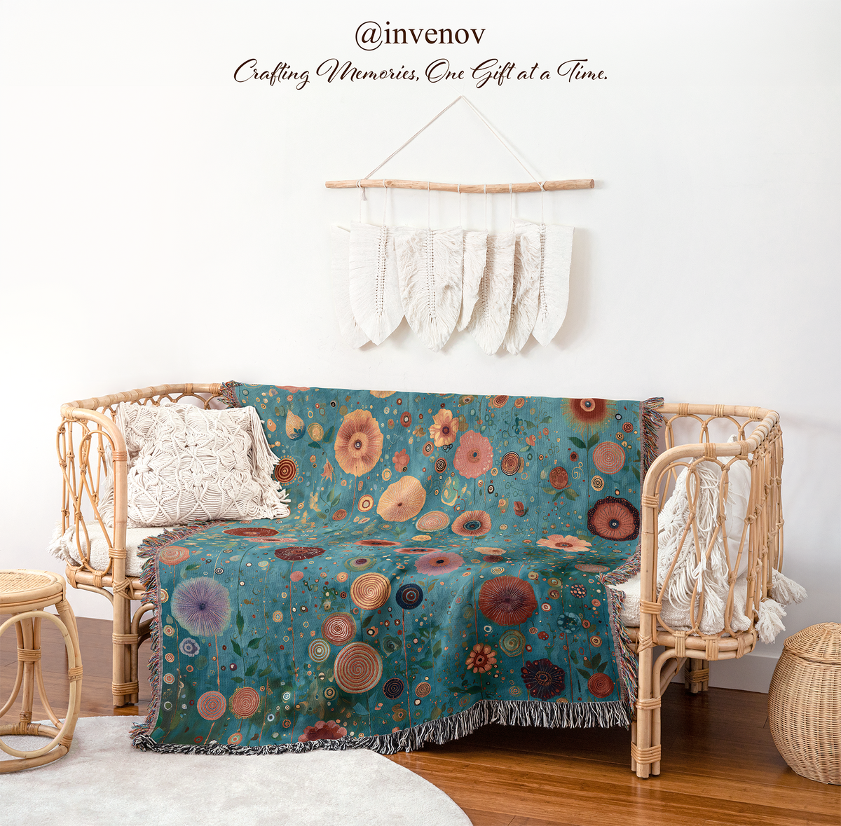 A teal bohemian throw blanket with a colorful floral and circular pattern is draped over a rattan daybed in a styled room setting. The scene includes a white wall with a macrame hanging, wooden flooring, and a small woven stool.