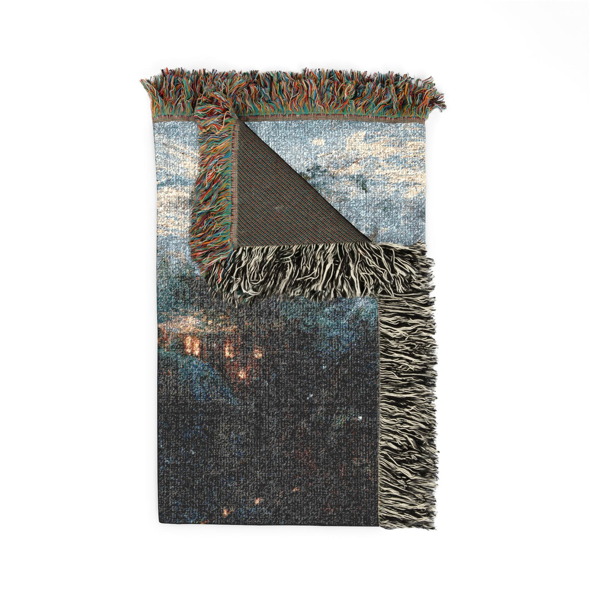 A folded woven blanket with multicolor fringe, featuring a distressed abstract landscape print in shades of blue, gray, and black with small warm light accents, shown on a white background