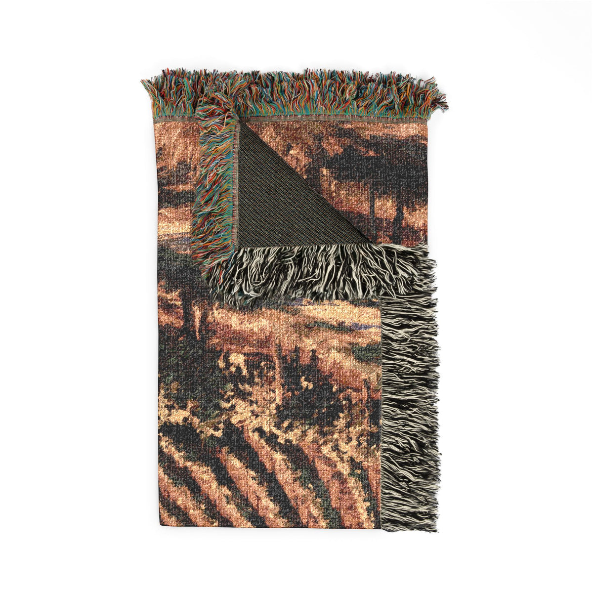 A rectangular woven throw blanket with a rustic landscape pattern in earthy tones, featuring fringed edges and a folded corner showing the reverse side, photographed on a solid white background.