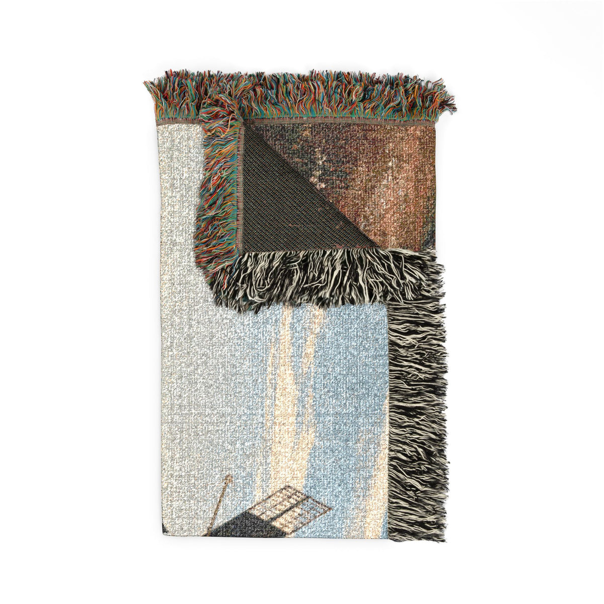 A folded woven throw blanket with multicolored fringe, featuring a light blue and cream landscape design with a dark brown textured section and a black-and-white fringed edge, shown on a plain white background.