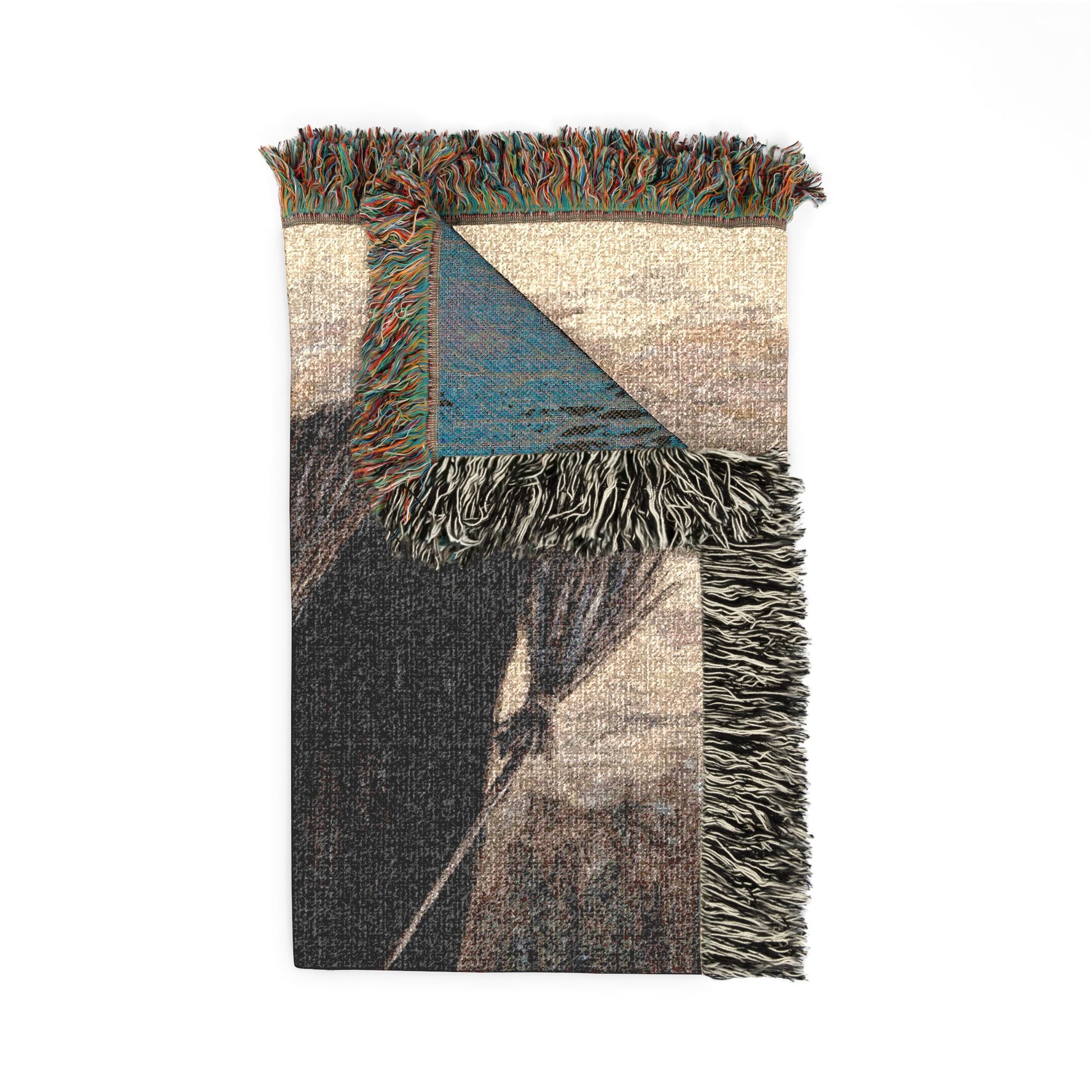 A folded woven throw blanket with fringed edges, featuring a geometric pattern in beige, charcoal gray, and a blue triangle, shown on a plain white background.