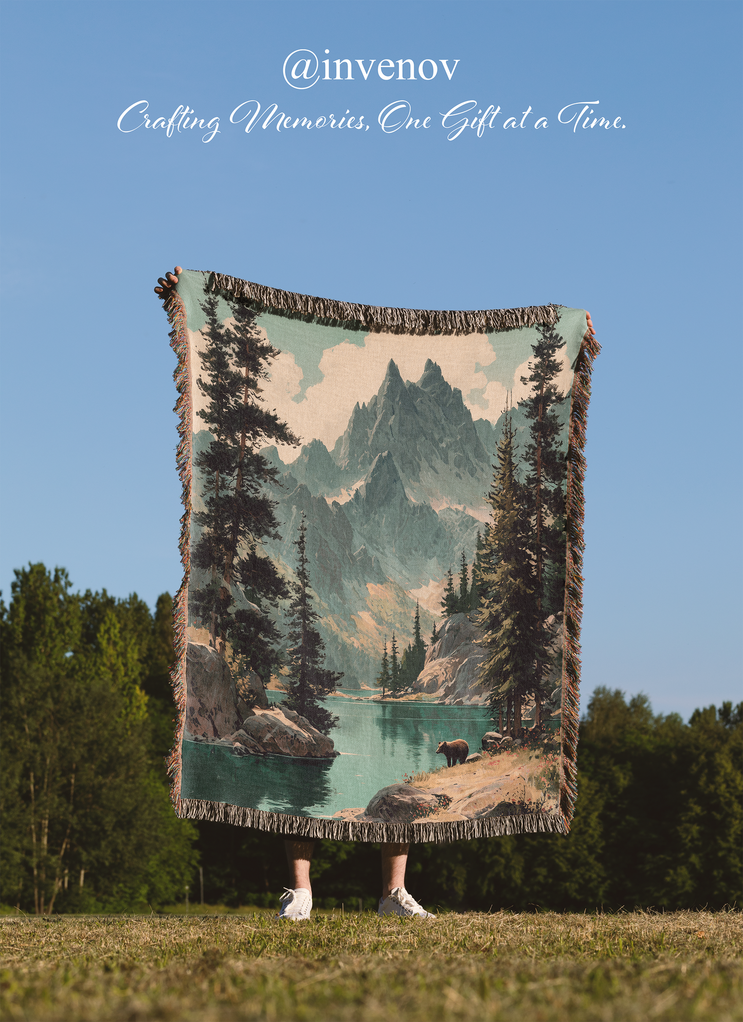 Person holding a scenic blanket with mountains and trees against a clear blue sky.
