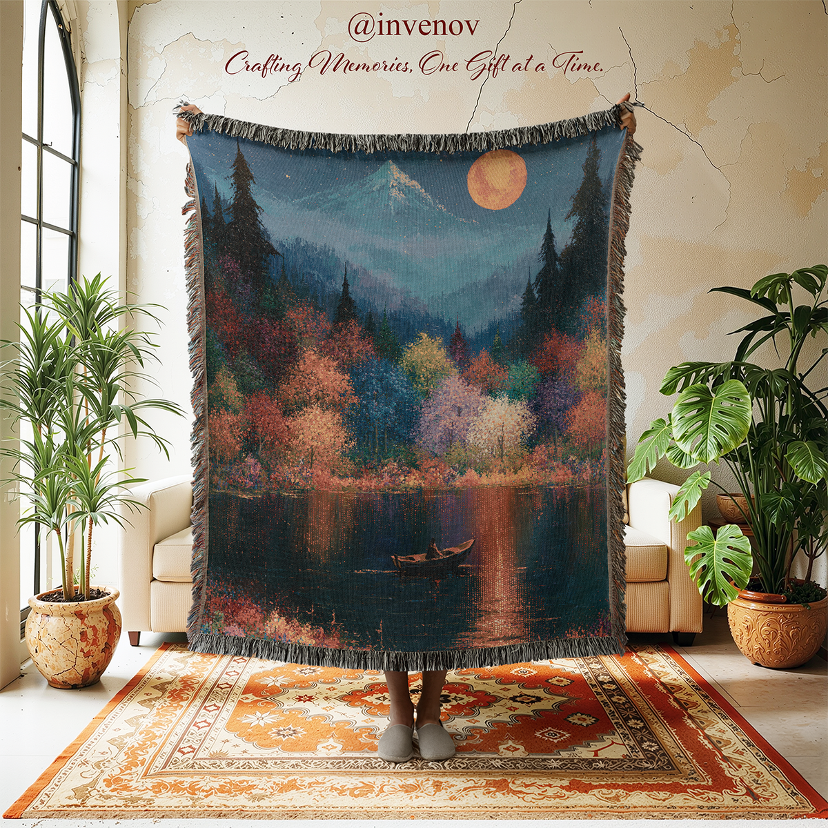 A large woven throw blanket with fringe is held up in a styled room. The blanket features a scenic landscape print of a lake, pine trees, mountains, and a small boat under a full moon.