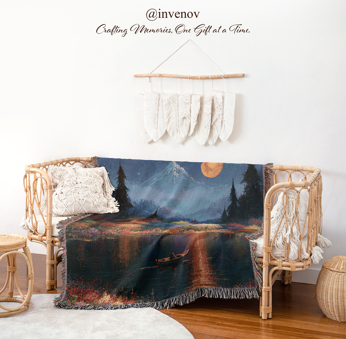 A woven throw blanket featuring a scenic night landscape print is draped over a rattan daybed in a styled room setting. The blanket depicts a mountain, lake, trees, and a small boat under a full moon.