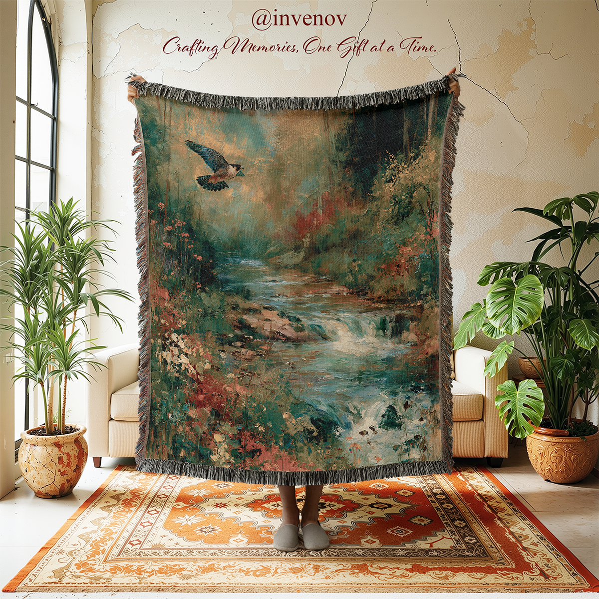 A person stands in a styled room holding up a large fringed throw blanket featuring a scenic landscape print of a river, trees, and a bird. The room has cream walls, a patterned rug, and potted plants.