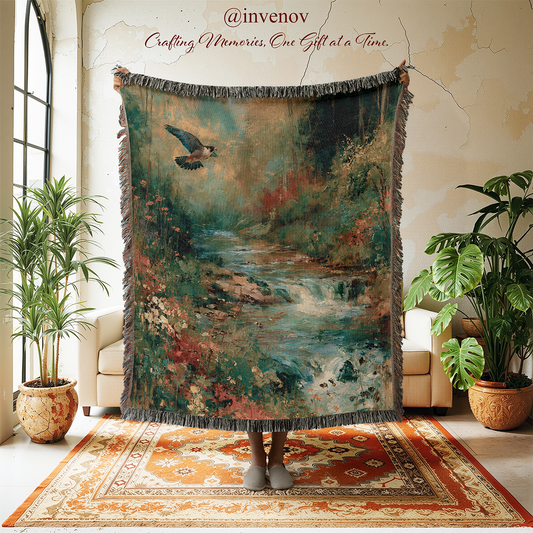 A person stands in a styled room holding up a large fringed throw blanket featuring a scenic landscape print of a river, trees, and a bird. The room has cream walls, a patterned rug, and potted plants.
