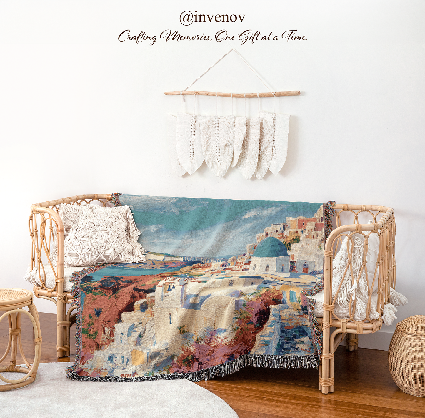 A woven throw blanket featuring a scenic Santorini coastal print with white buildings, blue domes, and a turquoise sea, styled on a rattan daybed in a boho room setting with a macrame wall hanging and decorative pillows.