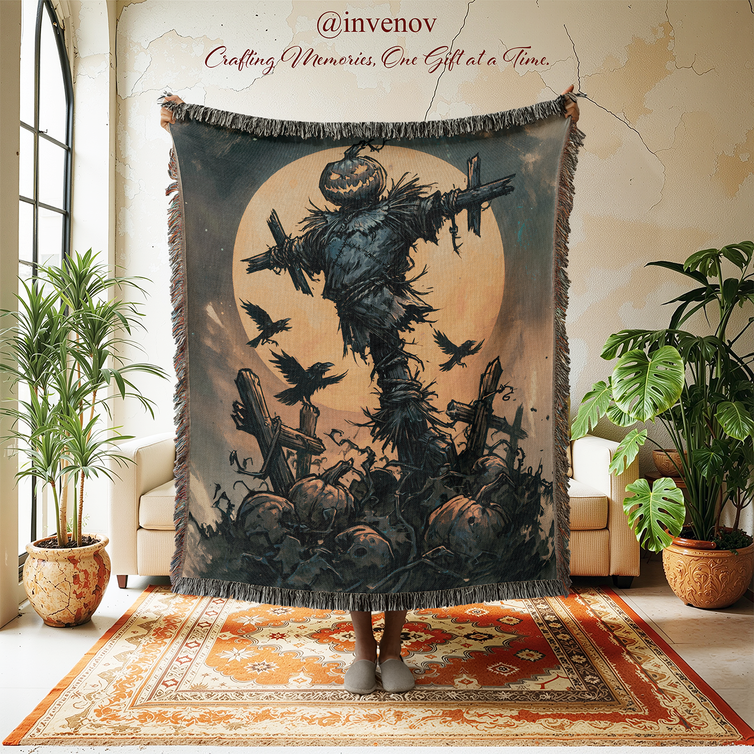 A person holds up a large woven throw blanket featuring a spooky Halloween scene with a scarecrow, pumpkins, crows, and a full moon. The blanket is shown in a styled room with plants, a rug, and cream-colored armchairs, with the brand name @invenov printed above.
