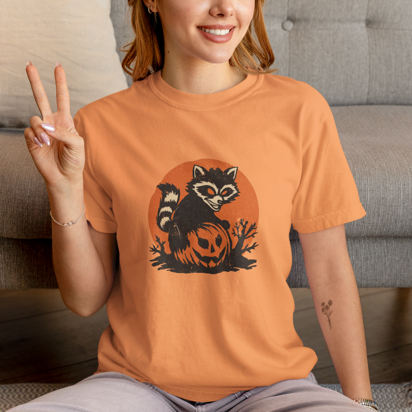 Person wearing an orange t-shirt with a raccoon and pumpkin design, sitting on a couch.