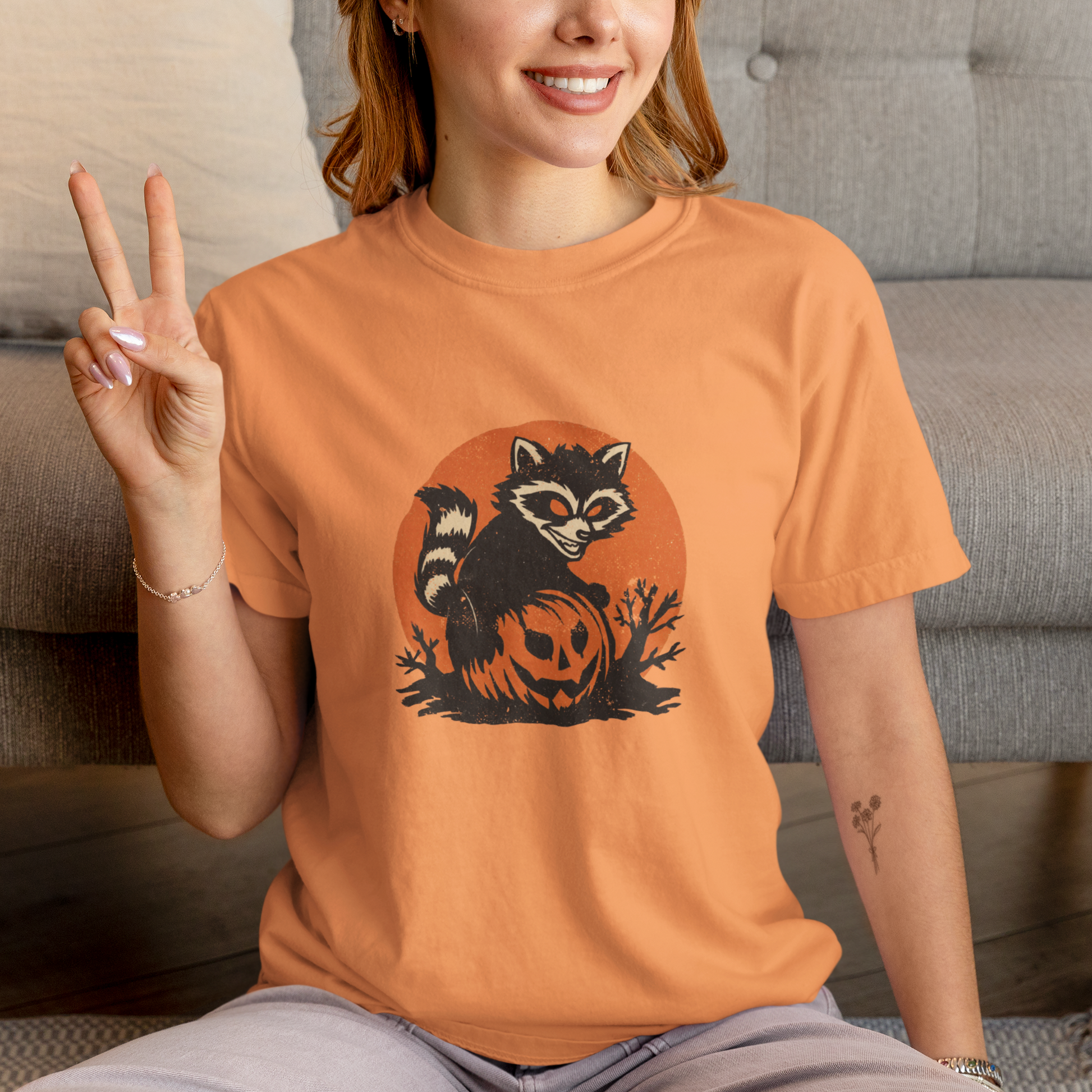 Person wearing an orange t-shirt with a raccoon and pumpkin design, sitting on a couch.