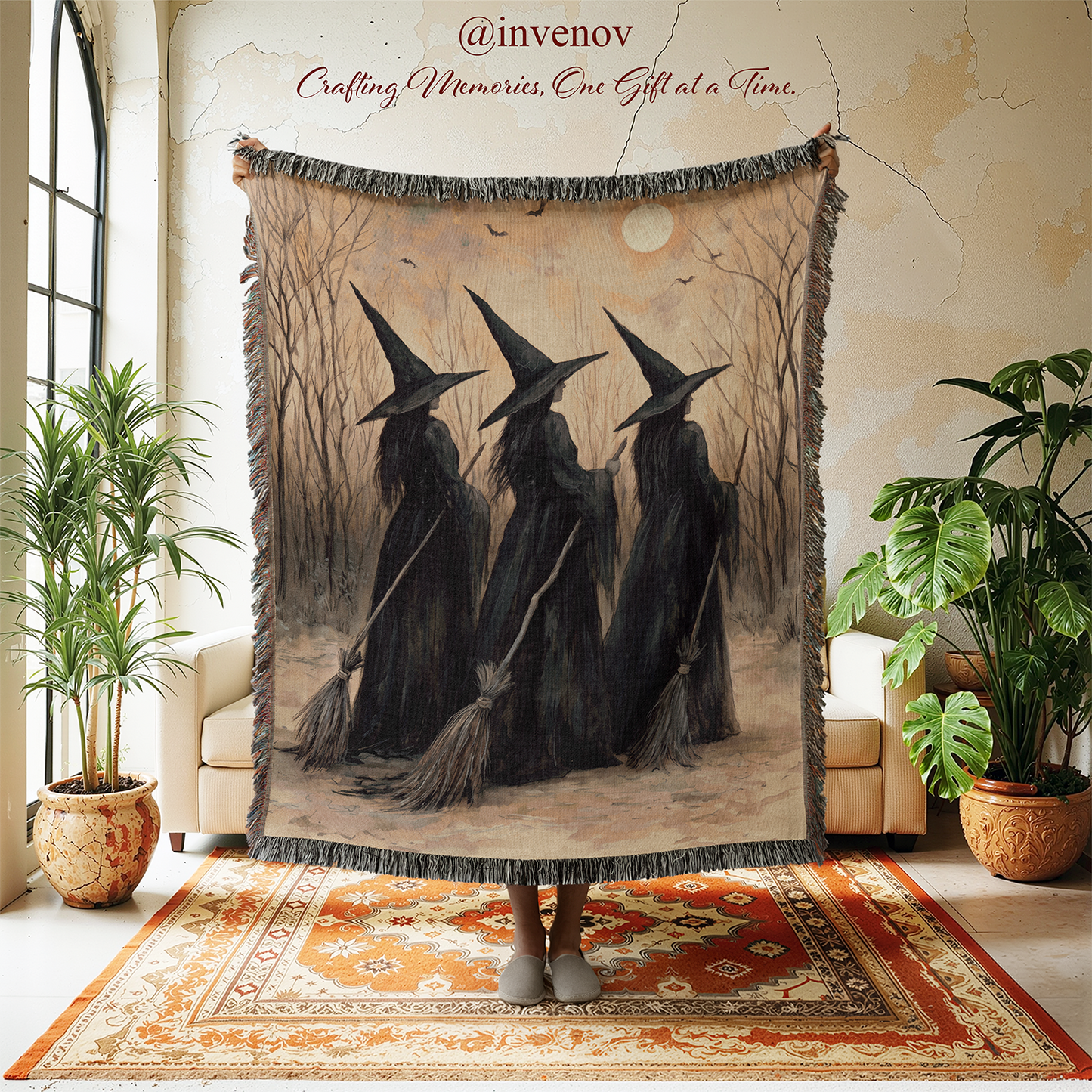 A person holds up a large fringed throw blanket in a styled room. The blanket features an illustration of three witches in black cloaks and pointed hats standing on broomsticks in a misty forest.