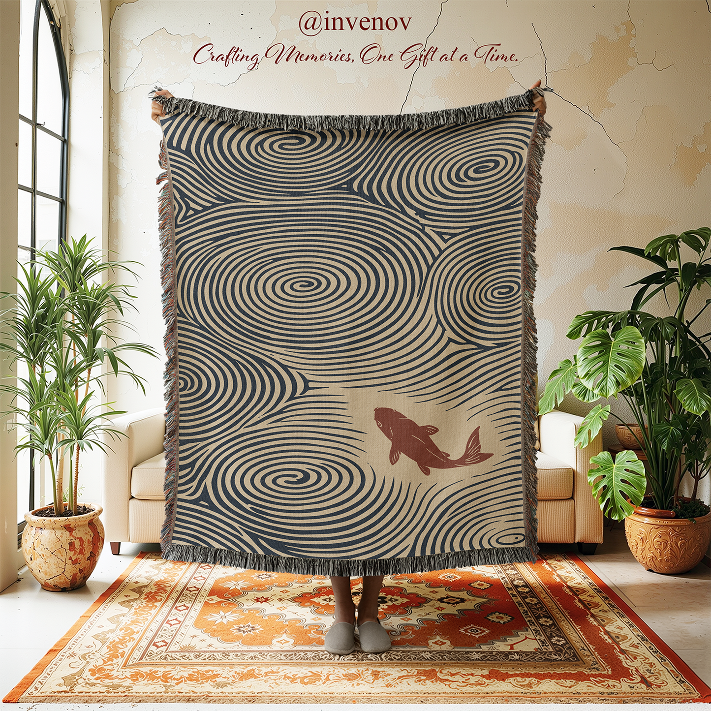 A person holds up a large fringed throw blanket in a styled room. The blanket has a beige background with dark navy swirling spiral patterns and a brown fish illustration near the lower right.