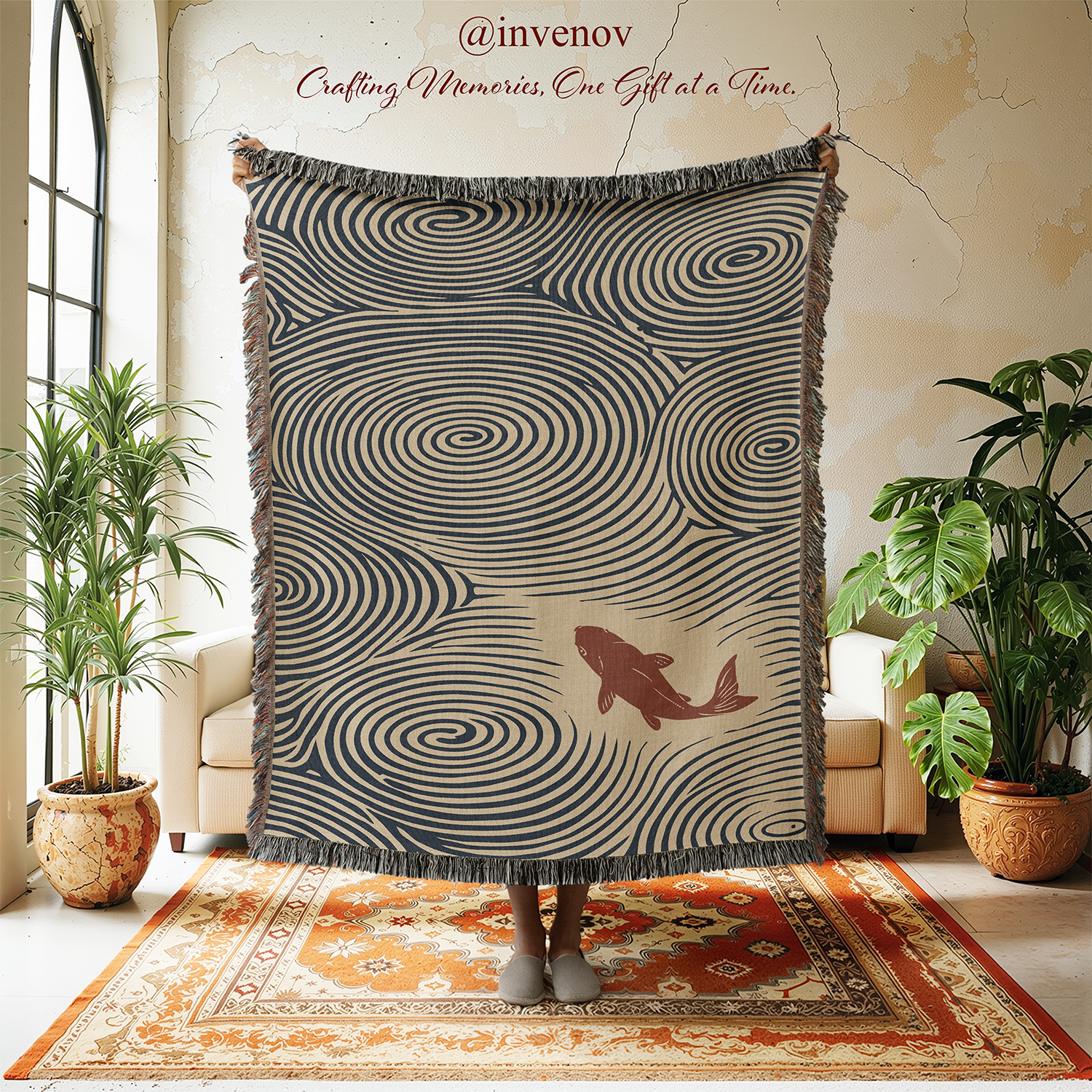 A person holds up a large fringed throw blanket in a styled room. The blanket has a beige background with dark navy swirling spiral patterns and a brown fish illustration near the lower right.
