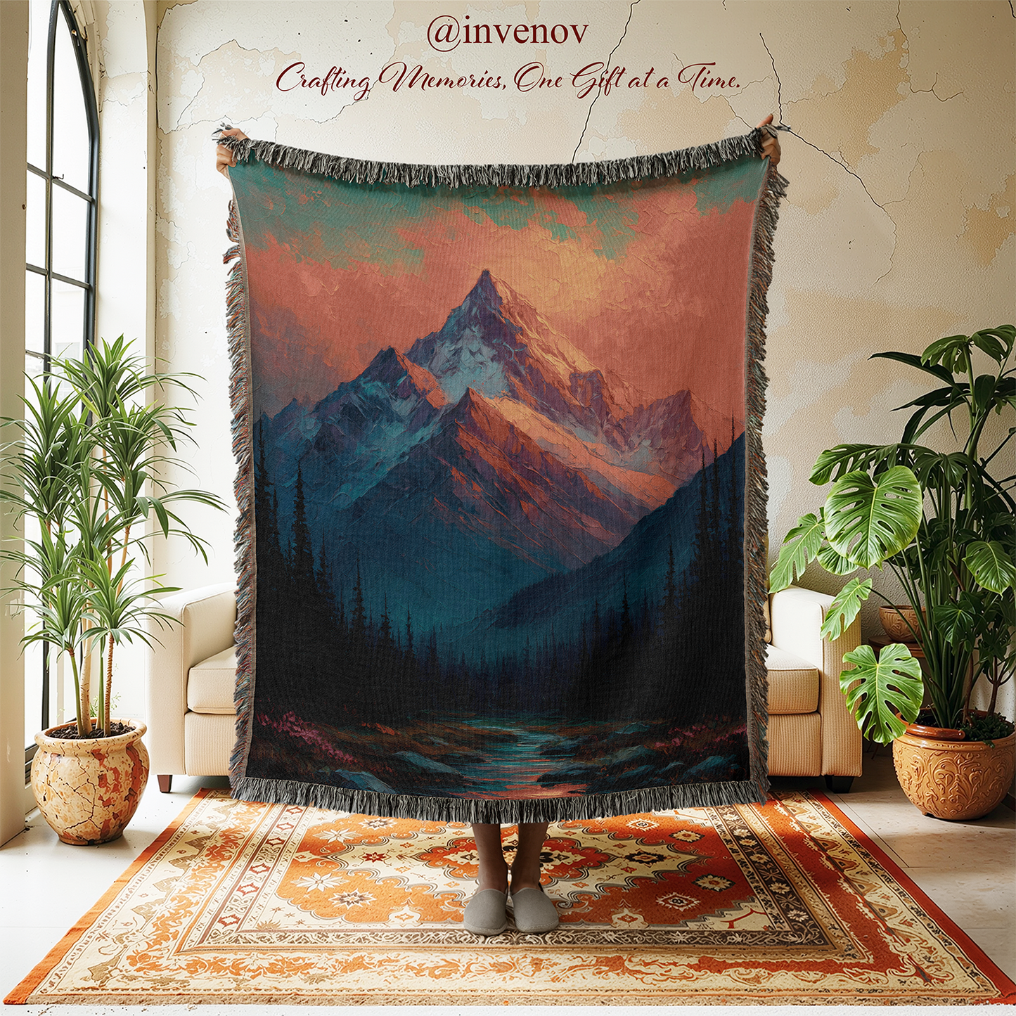 A large woven throw blanket with dark gray fringe is held up in a living room setting. The blanket features a scenic mountain landscape print with blue, teal, and orange tones.